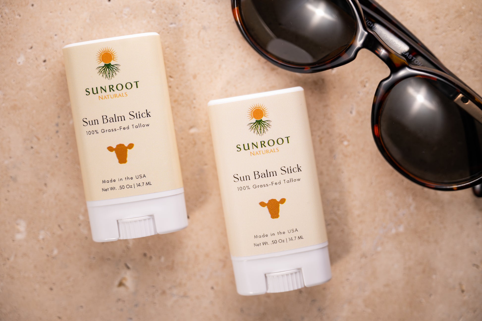 Two Sunroot Sun Balm Stick sunscreen sticks displayed beside sunglasses in a travel-inspired flat lay.